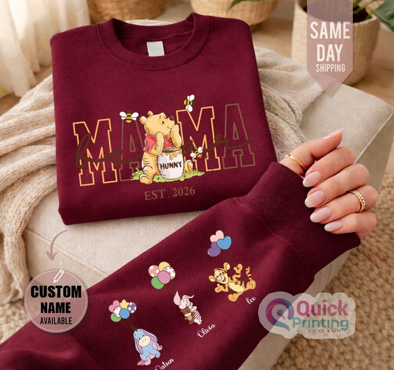 Puede incluir: Sudadera burdeos con un dise&ntilde;o de Winnie the Pooh, la palabra "MAMA" y "EST. 2026". La manga muestra personajes de dibujos animados con nombres. Tambi&eacute;n es visible el texto "CUSTOM NAME AVAILABLE".