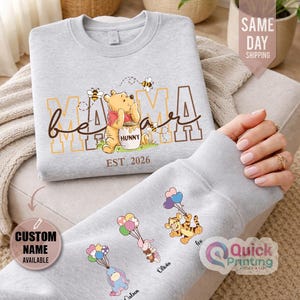 Puede incluir: Una sudadera gris claro con un dise&ntilde;o de Winnie the Pooh, con las palabras "Mama Bear" y "EST. 2026". La manga muestra personajes de dibujos animados con globos y nombres personalizados. Es una prenda de vestir c&oacute;moda.