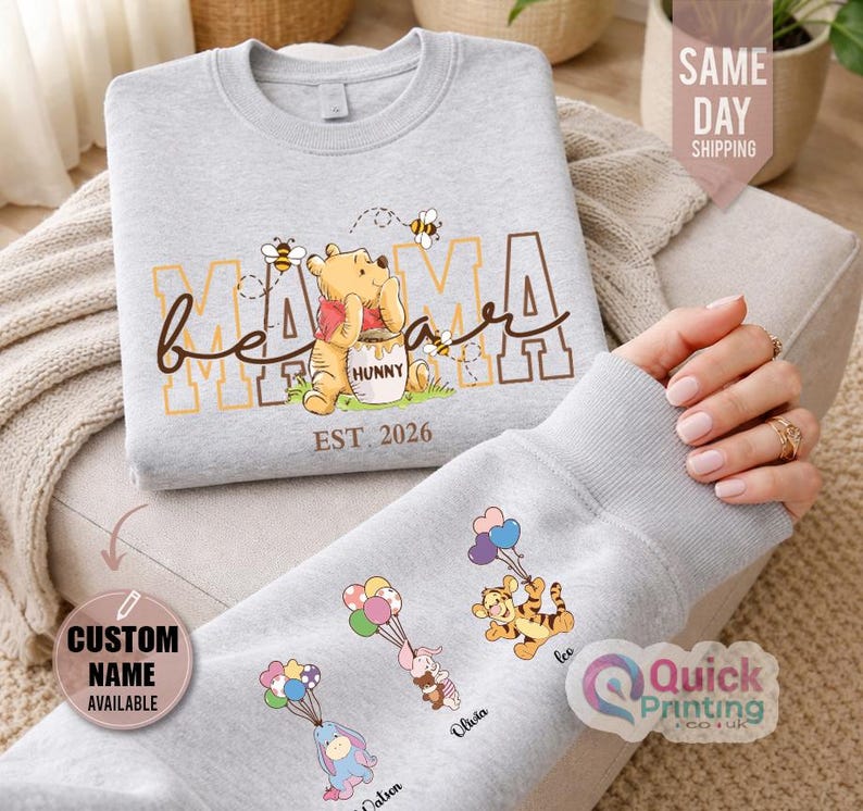 Puede incluir: Una sudadera gris claro con un dise&ntilde;o de Winnie the Pooh, con las palabras "Mama Bear" y "EST. 2026". La manga muestra personajes de dibujos animados con globos y nombres personalizados. Es una prenda de vestir c&oacute;moda.