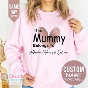 May include: A light pink sweatshirt with the text "This Mummy Belongs To Archie, Henry & Oliver" in black script. A rose gold heart shape is above the word "Mummy". A circular graphic says "Custom Name Available".
