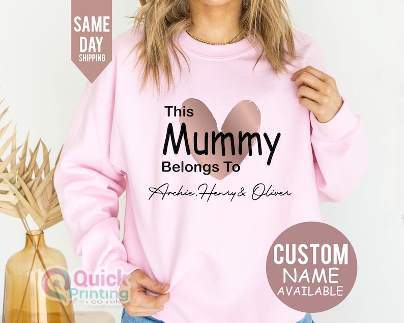 May include: A light pink sweatshirt with the text "This Mummy Belongs To Archie, Henry & Oliver" in black script. A rose gold heart shape is above the word "Mummy". A circular graphic says "Custom Name Available".