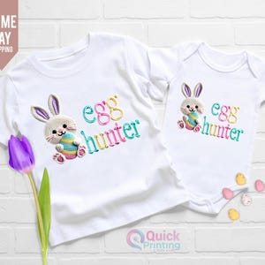 Op de afbeelding: Witte t-shirt en romper set met een geborduurde paashaas met een ei. De tekst "egg hunter" is in kleur geborduurd. Een paarse tulp staat in de linker benedenhoek. De achtergrond is een witte bakstenen muur.