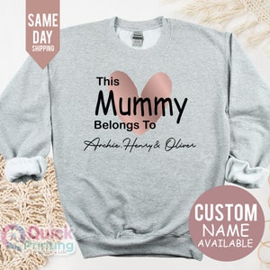 May include: A light gray sweatshirt with the text "This Mummy Belongs To Archie, Henry & Oliver" in black, with a pink heart design. The words "Custom Name Available" are also visible.