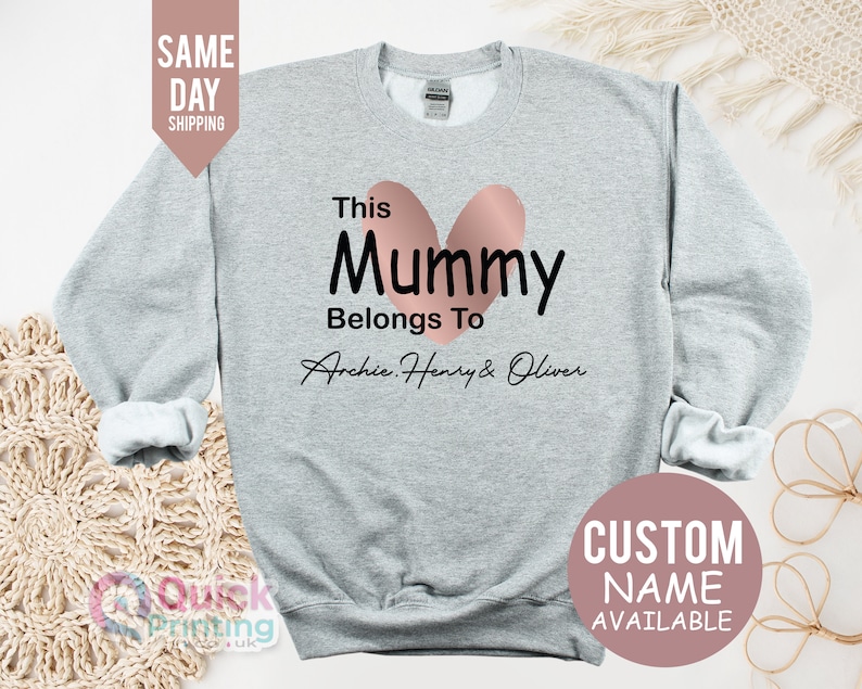 May include: A light gray sweatshirt with the text "This Mummy Belongs To Archie, Henry & Oliver" in black, with a pink heart design. The words "Custom Name Available" are also visible.