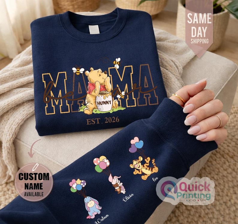 Puede incluir: Sudadera azul marino con dise&ntilde;o de Winnie the Pooh, la palabra "MAMA" y "EST. 2026". La manga muestra personajes de dibujos animados con globos y nombres. Una sudadera c&oacute;moda e informal.