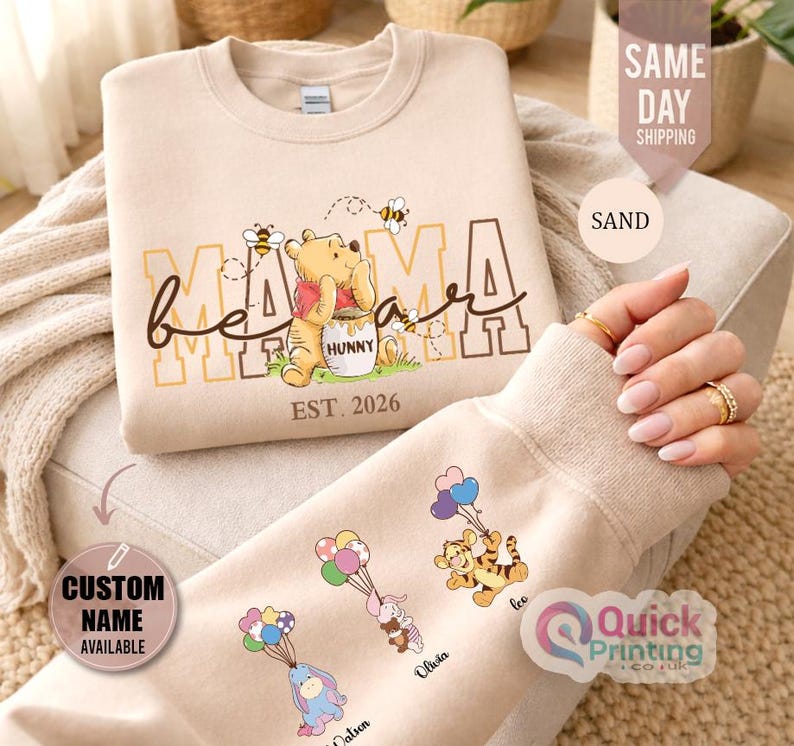 Puede incluir: Sudadera color arena con la frase "Mama Bear" y un gr&aacute;fico de Winnie the Pooh. La manga presenta personajes de dibujos animados con globos. El texto "Custom Name Available" es visible.