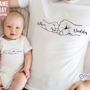 May include: White baby bodysuit and t-shirt with a black outline of two fists bumping, one smaller than the other. The text "Arthur" and "Daddy" are written next to the fists.