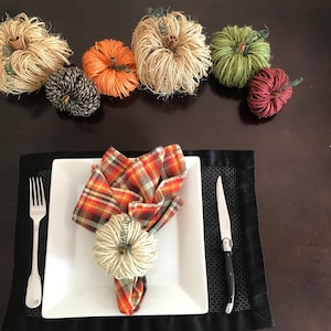 May include: A fall-themed table setting with a plaid napkin ring, a white plate, a fork, and a butter knife. The table is decorated with small, handmade yarn pumpkins in various colors, including orange, green, brown, and red.