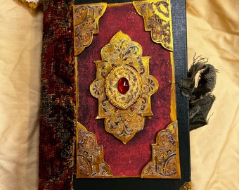May include: A rectangular journal with a black cover, featuring a central red panel adorned with intricate gold filigree and a single red gemstone. The spine is wrapped in textured dark red fabric, and the corners have gold metal accents.