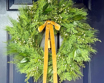 May include: A circular wreath composed of green pine needles, cedar sprigs, and small white berries. A long, golden-brown velvet ribbon is tied in a bow at the top centre, with two long tails hanging down. A small brass bell hangs behind the ribbon.