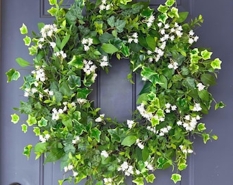 May include: A circular green wreath with white flowers and ivy leaves is displayed on a blue door. The wreath is made of artificial plants. The door features a black handle and a digital lock.