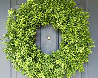 May include: A bright green, leafy wreath hangs on a grey door. The wreath is circular and densely packed with foliage. The door has a simple panel design and a dark-coloured doorknob. The overall effect is fresh and inviting.