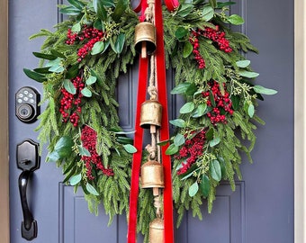 May include: A Christmas wreath featuring green foliage, red berries, and three brass bells suspended by a red ribbon. The wreath is displayed on a blue door, a traditional festive decoration for the holiday season.