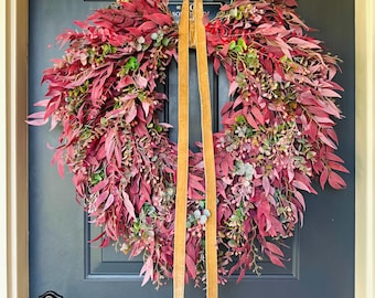 May include: A full wreath with burgundy and green leaves, and small berries. The wreath is suspended by a gold-coloured hanger and tan ribbons, creating a welcoming display for a front door.