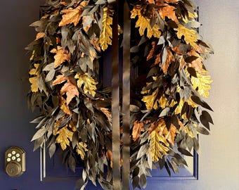 May include: A circular wreath composed of dark black leaves and metallic gold oak leaves, featuring a long black satin ribbon hanging from the top centre. The wreath is mounted on a dark blue door with a visible electronic keypad lock and handle.
