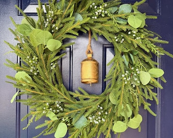 May include: A circular wreath constructed from green pine needles and eucalyptus leaves, accented with small white berries. A single, weathered gold-toned metal bell hangs from a jute rope in the centre of the wreath, which is suspended by a gold hook.
