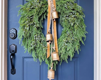 May include: A Christmas wreath made of green foliage and small blue-green berries, with gold bells suspended from a tan ribbon. The wreath hangs on a navy blue door, a festive seasonal decoration.