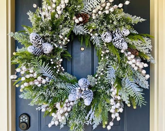 May include: A Christmas wreath featuring green and frosted white foliage, white berries, and pinecones. The wreath is displayed on a dark blue door, creating a festive holiday decoration for the home.