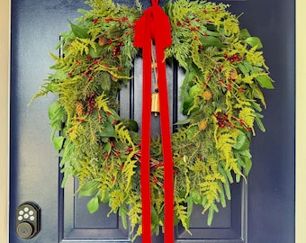 May include: A circular wreath made of green pine needles, fern-like foliage, and small red berries, accented with brown pinecones. A long, bright red velvet ribbon hangs from the top, featuring a small gold-coloured metal bell at the centre.