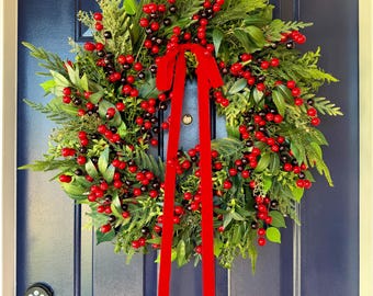 May include: A circular wreath composed of lush green foliage, including pine-like needles and broad leaves, densely accented with clusters of bright red and dark berries. A long, vibrant red velvet ribbon is tied at the top, hanging down the centre.