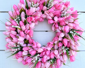 May include: A circular wreath made of pink tulip flowers, with small white flowers and green leaves. The wreath is densely packed with the tulip blooms, creating a full and vibrant appearance. The background is a white wooden surface.