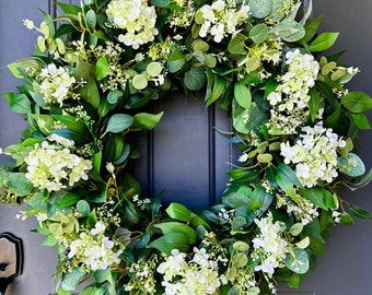 May include: A circular wreath composed of lush green foliage and white hydrangea flowers. The wreath is adorned with various types of leaves and small white blossoms, creating a full and vibrant appearance. The wreath is displayed on a dark gray door.