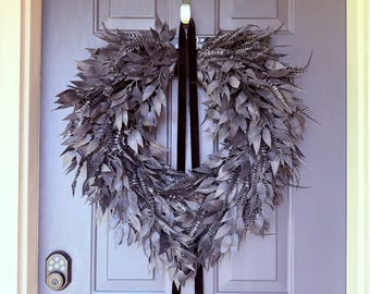 May include: A grey leaf wreath hangs on a grey door. The wreath is made of layered leaves and has a dark ribbon. The door has a smart lock and a silver handle. The wreath is a decorative item.