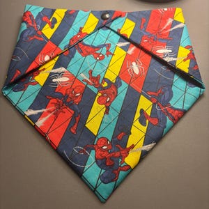 以下が含まれることがあります： 赤と青のスーツを着たスパイダーマン、白いクモのアイコン、細い黒いクモの巣の線が繰り返される模様の三角形のペット用バンダナ。生地の背景は、黄色、水色、濃い青の斜めのストライプで構成されており、黒いスナップボタンが付いています。