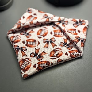 May include: A folded fabric bandana featuring a repeating pattern of brown American footballs and brown bows on a cream-colored background. The material has a textured surface and is folded into a triangular shape with visible edges.