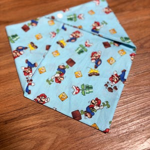 以下が含まれることがあります： マリオのキャラクター、緑の土管、ハテナブロック、パックンフラワーが繰り返し描かれた、水色の三角形の布製バンダナ。布は先端が尖るように折りたたまれており、ゲームの様々なアイコンが表示されています。