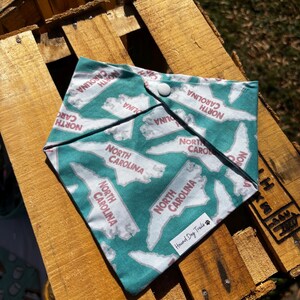 May include: A teal blue bandana with a white outline of the state of North Carolina repeated across the fabric. The text "North Carolina" is printed in pink on each state outline. The bandana has a snap closure and the text "Hound Dog Trails" is printed in white on a paw print.