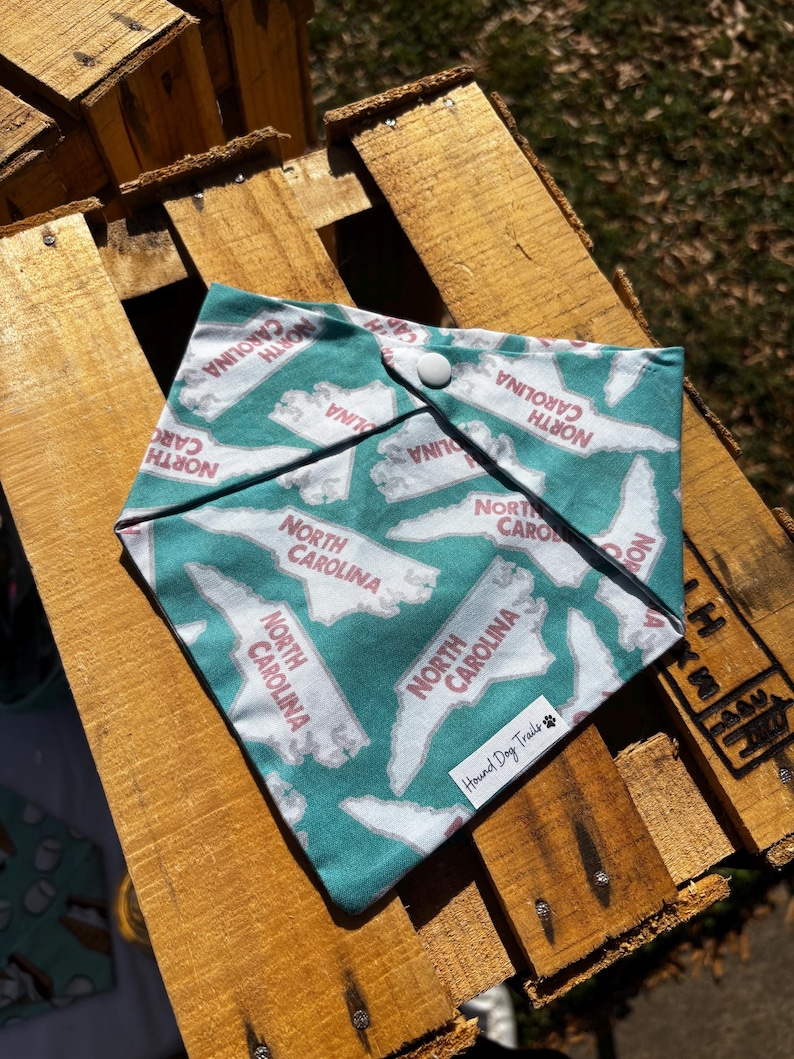 May include: A teal blue bandana with a white outline of the state of North Carolina repeated across the fabric. The text "North Carolina" is printed in pink on each state outline. The bandana has a snap closure and the text "Hound Dog Trails" is printed in white on a paw print.
