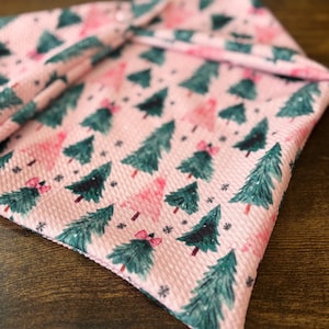 May include: A triangular fabric bandana featuring a light pink background with a repeating pattern of dark green and light pink Christmas trees, small black snowflakes, and tiny pink bows attached to some of the tree tops.