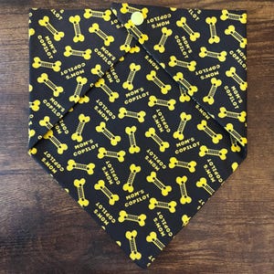 May include: A black triangular pet bandana featuring a repeating yellow pattern of dog bones and the text MOM'S COPILOT. The fabric is secured with a single light yellow snap button at the top corner, creating a folded, layered appearance.