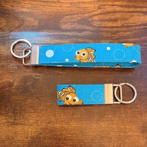 May include: Two blue fabric keychains with a cartoon fish pattern. The keychains have silver metal hardware and a loop for keys. The fabric has a light blue background with white circles and orange and yellow fish.