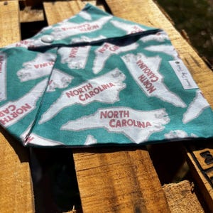 May include: A teal blue bandana with a white outline of the state of North Carolina repeated across the fabric. The state outline is filled in with red text that says "North Carolina". The bandana has a snap closure.