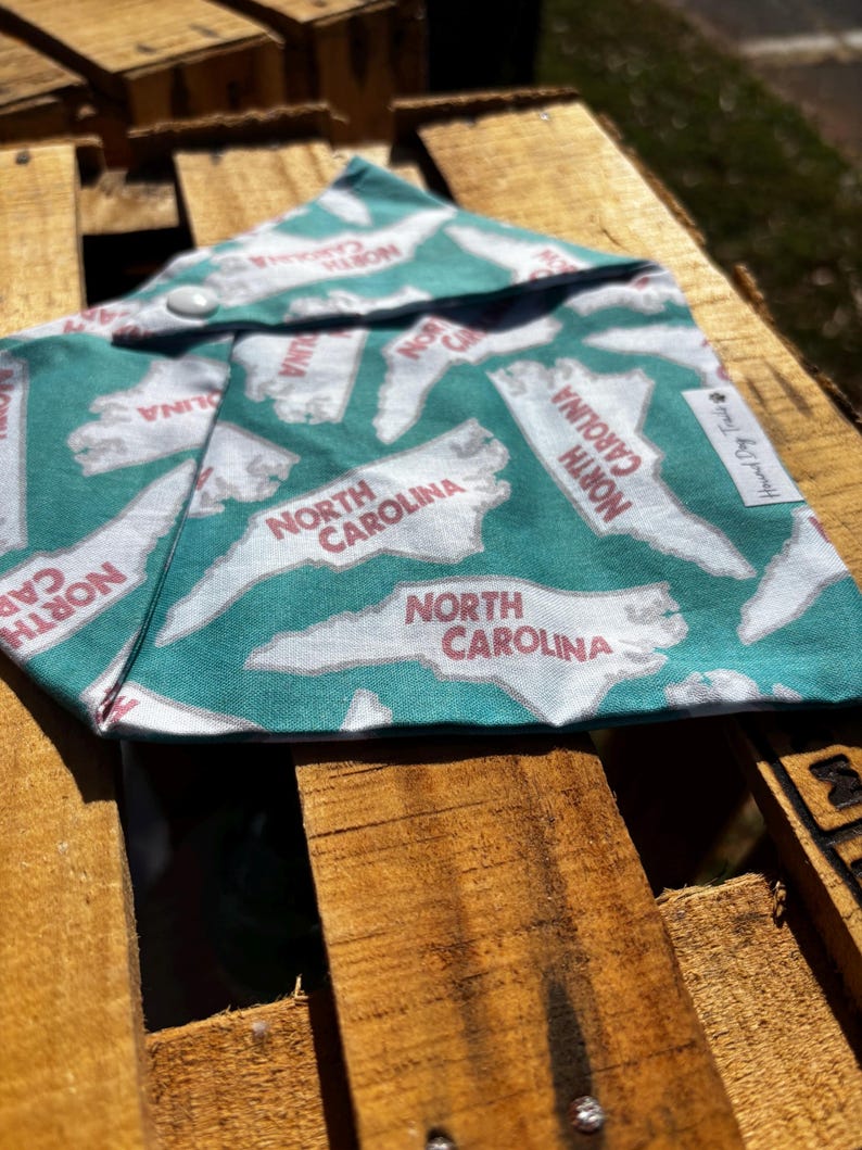 May include: A teal blue bandana with a white outline of the state of North Carolina repeated across the fabric. The state outline is filled in with red text that says "North Carolina". The bandana has a snap closure.