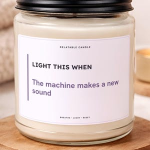 May include: A clear glass candle jar with a black metal lid, filled with off-white wax. The white label features the text RELATABLE CANDLE, LIGHT THIS WHEN, The machine makes a new sound, and BREATHE LIGHT RESET in black and purple ink.