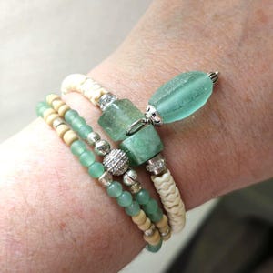 May include: A multi-strand bracelet featuring light green frosted beads, cream-colored textured beads, and square green stone accents. The piece includes a dangling seafoam green glass charm, silver-toned metal spacers, and small clear crystals.