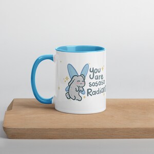 May include: White ceramic mug with a light blue interior and handle. The mug features a cartoon bunny with blue fairy wings and the text "You are so so so Radiant!" on the front. The mug is sitting on a wooden surface.