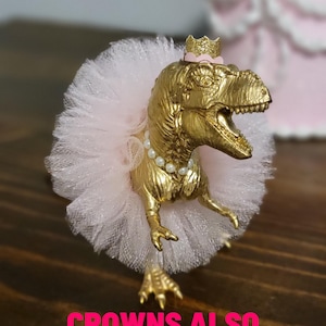 May include: A gold-colored Tyrannosaurus Rex figurine wearing a pink tutu, pearl necklace, and a small gold crown. The text "CROWNS ALSO AVAILABLE" is displayed in pink. The figurine is likely a cake topper or decorative item.