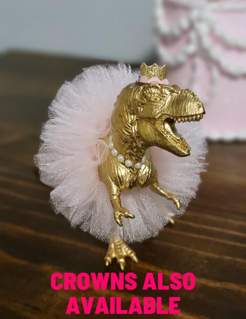 May include: A gold-colored Tyrannosaurus Rex figurine wearing a pink tutu, pearl necklace, and a small gold crown. The text "CROWNS ALSO AVAILABLE" is displayed in pink. The figurine is likely a cake topper or decorative item.
