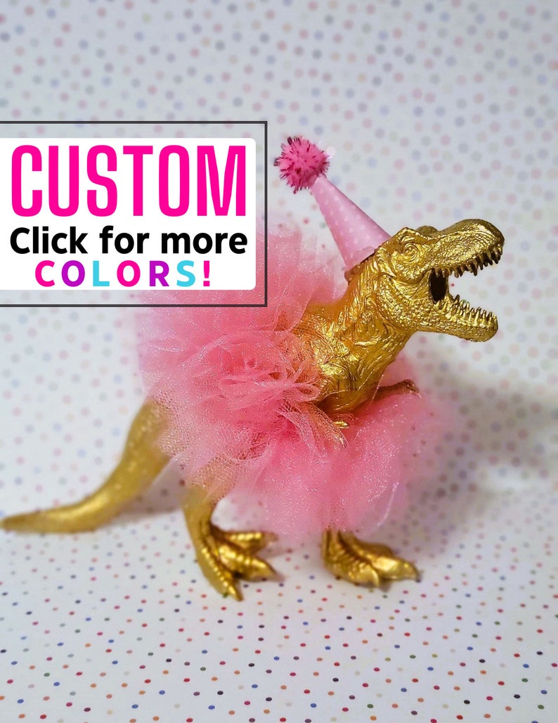 May include: A gold-painted Tyrannosaurus Rex figurine wearing a pink tutu and party hat with a pom-pom. The image includes the text "CUSTOM Click for more COLORS!" in a white box with pink and blue text.