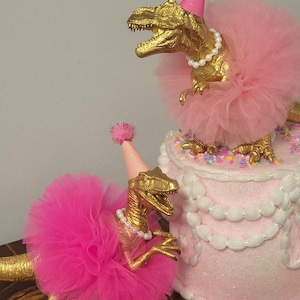 May include: Two gold-colored Tyrannosaurus Rex figurines wearing pink tutus, party hats, and pearl necklaces. One dinosaur is perched on a pink cake decorated with white frosting and sprinkles. The other dinosaur is on a wooden surface.