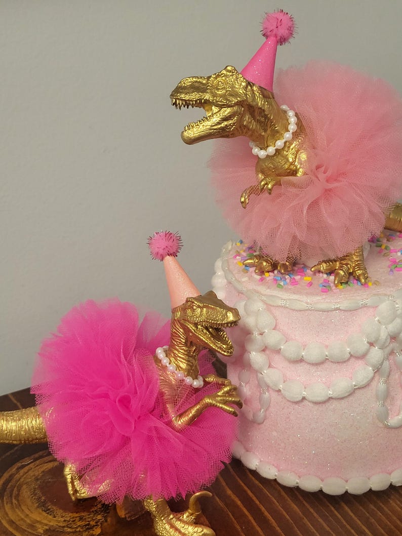 May include: Two gold-colored Tyrannosaurus Rex figurines wearing pink tutus, party hats, and pearl necklaces. One dinosaur is perched on a pink cake decorated with white frosting and sprinkles. The other dinosaur is on a wooden surface.