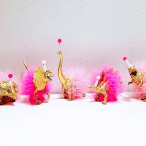 May include: Five gold-painted toy dinosaurs wearing pink tutus and party hats with pink pom-poms. The dinosaurs are arranged in a row, creating a whimsical and festive scene.