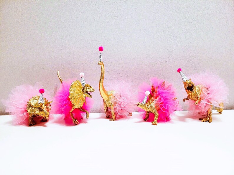 May include: Five gold-painted toy dinosaurs wearing pink tutus and party hats with pink pom-poms. The dinosaurs are arranged in a row, creating a whimsical and festive scene.