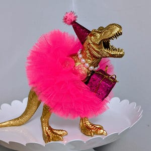 May include: A gold-colored Tyrannosaurus Rex figurine wearing a pink tutu, party hat, and pearl necklace. The dinosaur holds a small, magenta gift box. The figurine is displayed on a white, scalloped-edge cake stand.