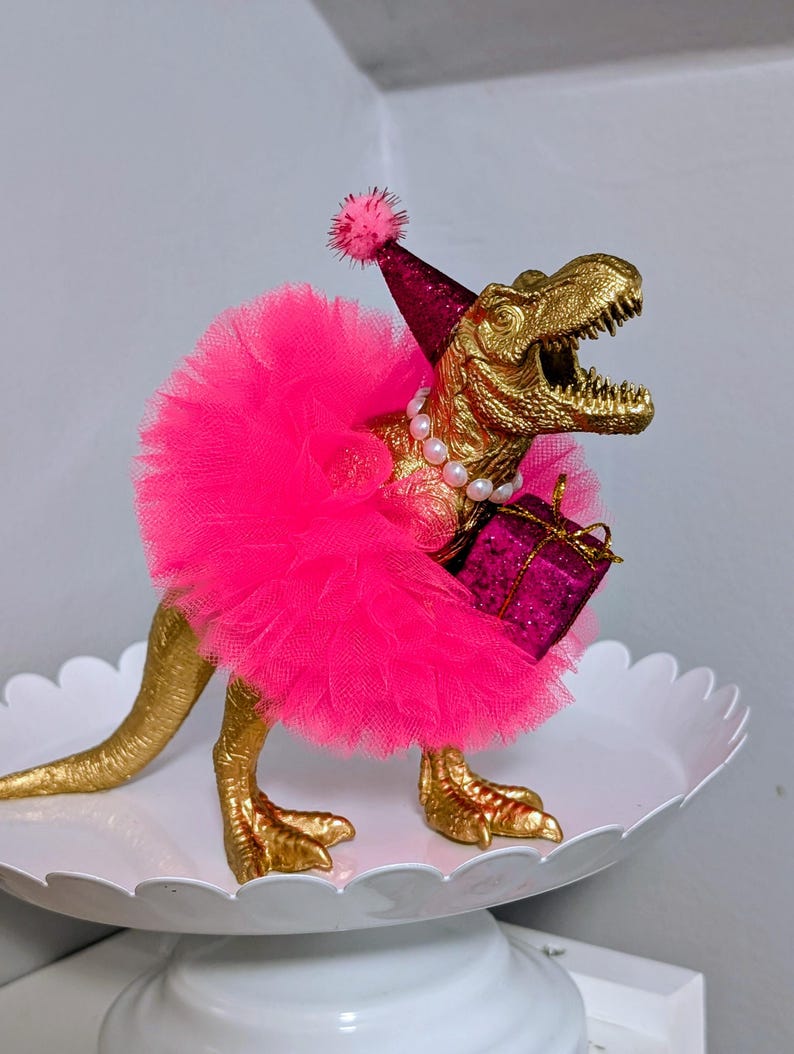 May include: A gold-colored Tyrannosaurus Rex figurine wearing a pink tutu, party hat, and pearl necklace. The dinosaur holds a small, magenta gift box. The figurine is displayed on a white, scalloped-edge cake stand.