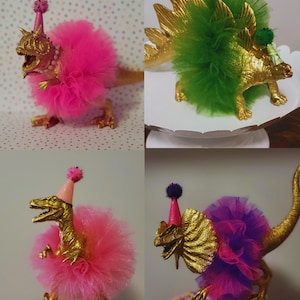 May include: Four gold-painted dinosaur figurines, each adorned with a colorful tutu and party hat. The text "DINO 5", "DINO 6", "DINO 7", and "DINO 8" are displayed in pink. The dinosaurs are cake toppers.